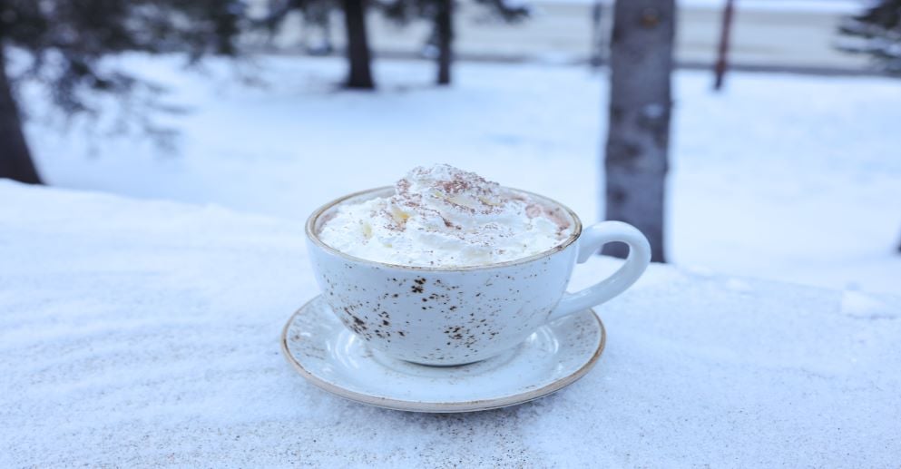 white mug filled with whipped cream topped drink sits on snow covered table outside