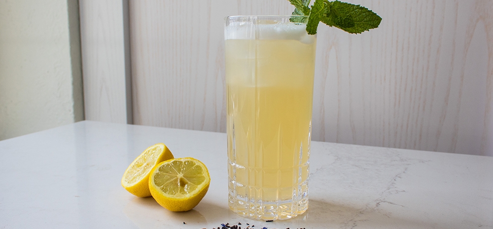 yellow coloured cocktail in a tall glass sits on table beside lemon halves