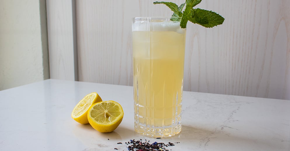 yellow coloured cocktail in a tall glass sits on table beside lemon halves
