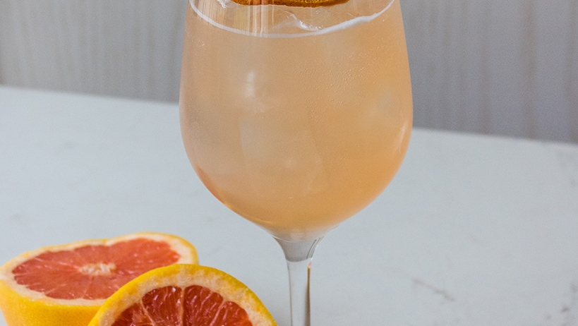 orange coloured cocktail in a wine glass sits on table beside grapefruit halves