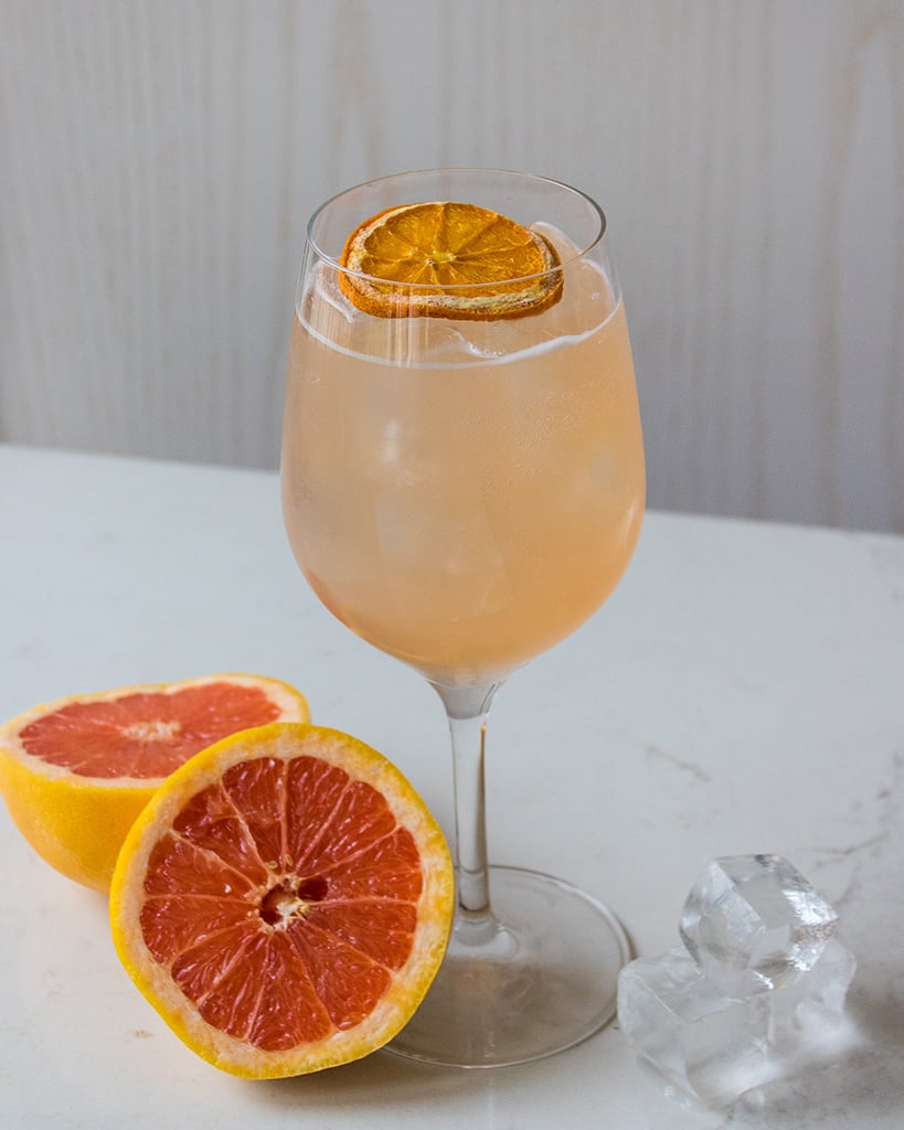 orange coloured cocktail in a wine glass sits on table beside grapefruit halves