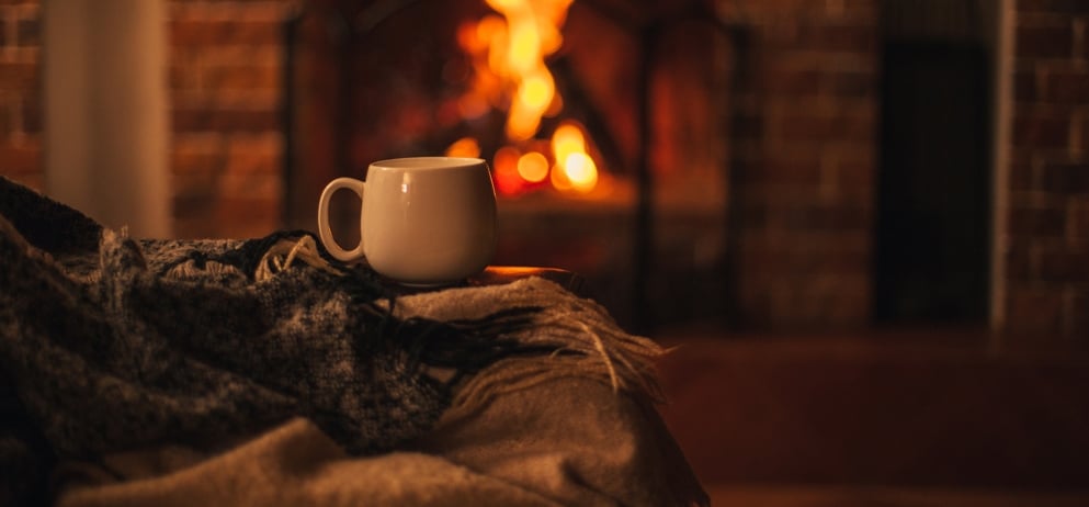 Mug with hot tea standing on a chair with woolen blanket in a cozy living room with fireplace. 