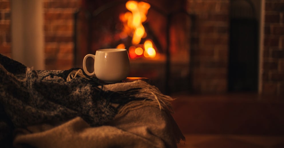 Mug with hot tea standing on a chair with woolen blanket in a cozy living room with fireplace. 