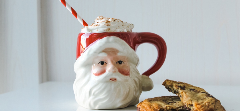 santa claus mug filled with whipped cream and red and white striped straw sits on table beside cookies