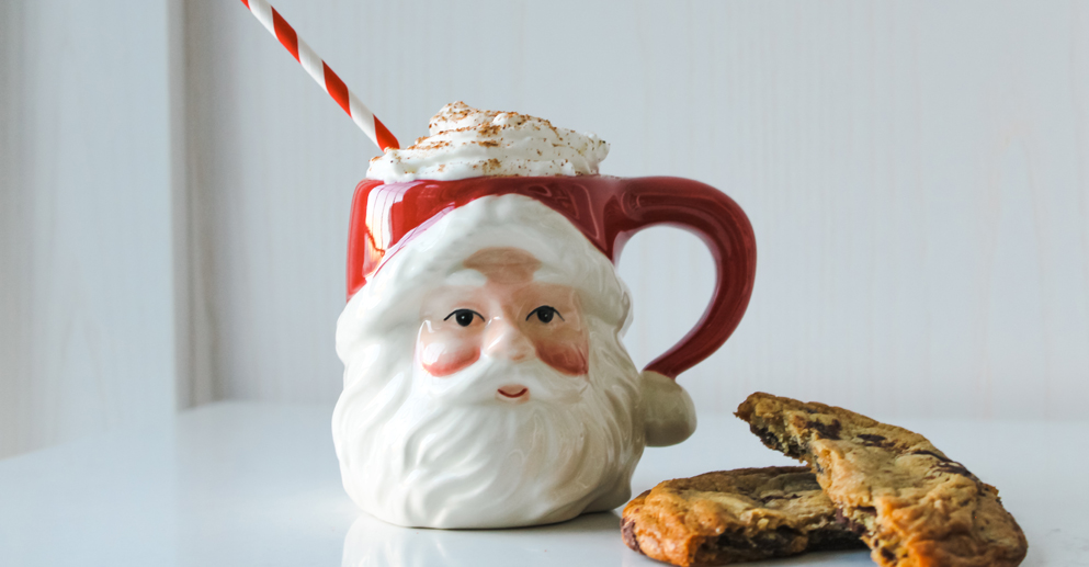 santa claus mug filled with whipped cream and red and white striped straw sits on table beside cookies