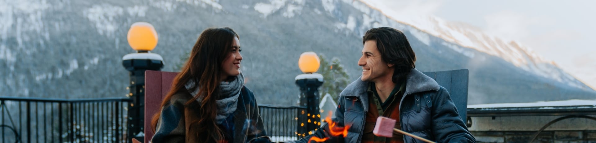 A couple holds hands and sits by a fire outdoors in the mountains while somebody off to the right is roasting a marshamallow.