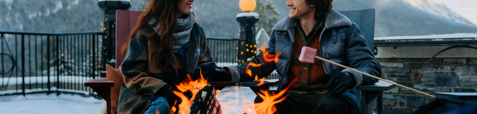 A couple holds hands and sits by a fire outdoors in the mountains while somebody off to the right is roasting a marshmallow.