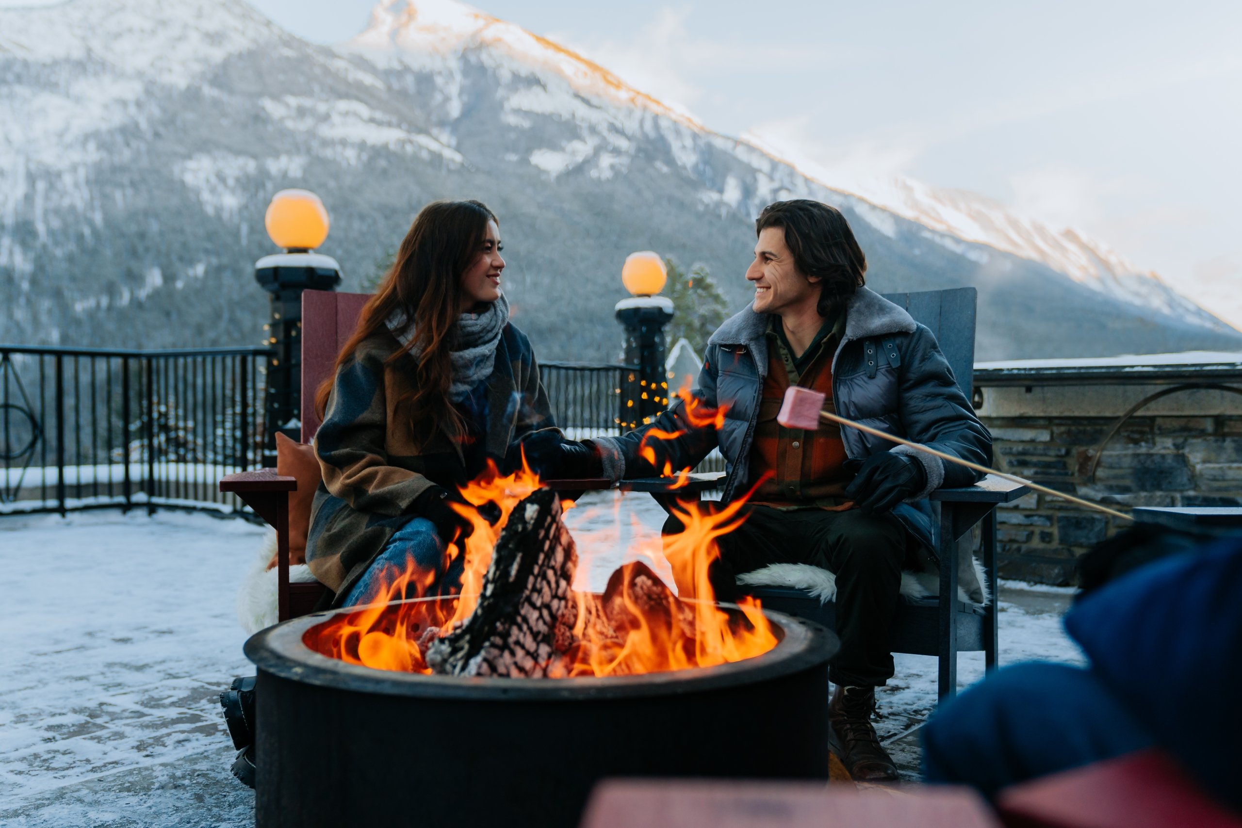 A couple holds hands and sits by a fire outdoors in the mountains while somebody off to the right is roasting a marshmallow.