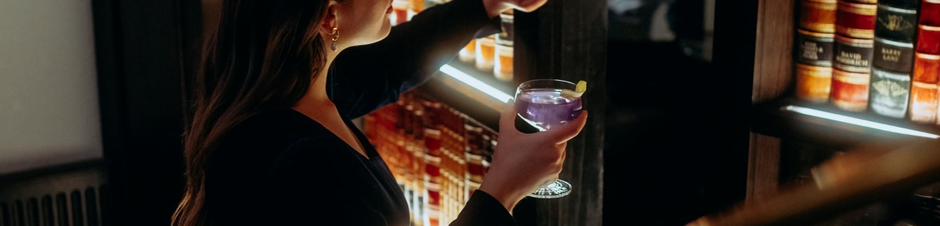 Woman peering from behind a secret bookcase door with a cocktail in her hand