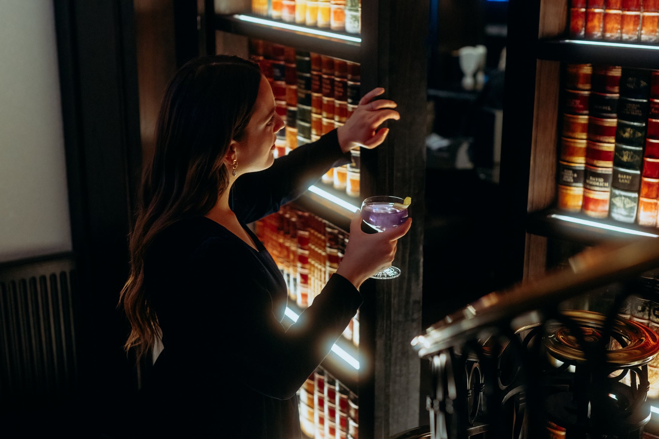 Woman peering from behind a secret bookcase door with a cocktail in her hand